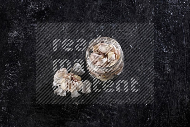 A glass jar full of garlic cloves and beside it a garlic bulb on a textured black flooring in different angles