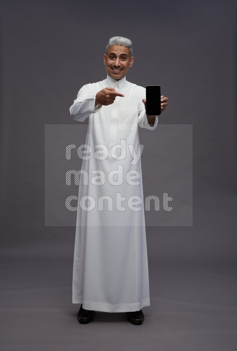 Saudi man wearing thob standing showing phone to camera on gray background
