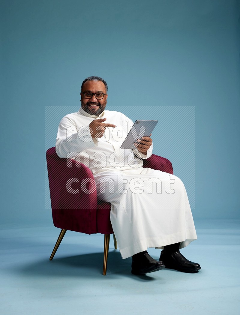 Saudi Man without shimag sitting on chair working on tablet on blue background