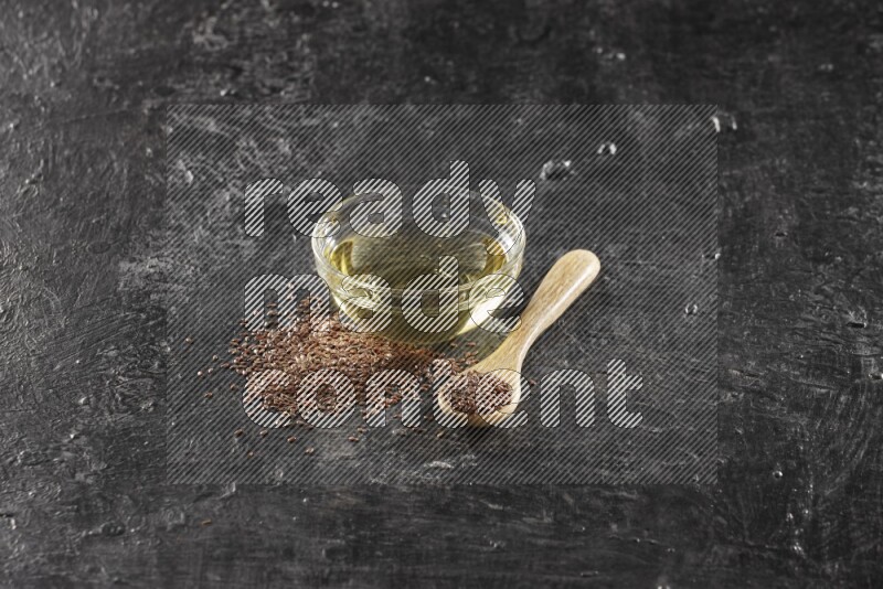 A glass bowl full of flaxseeds oil and wooden spoon full of flaxseeds with seeds spread on a textured black flooring