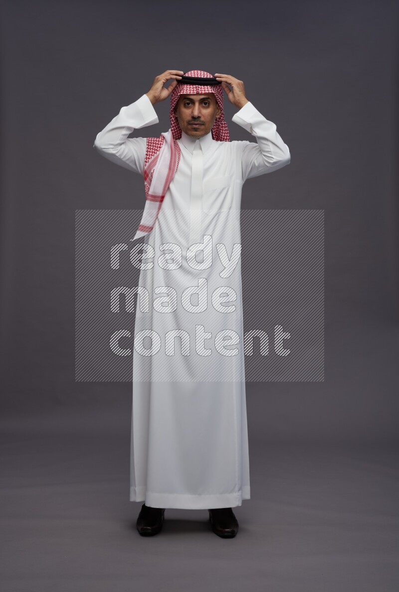 Saudi man wearing thob and shomag standing hands behind head on gray background