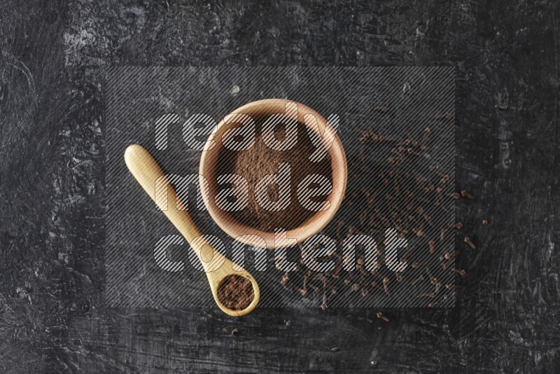 A wooden bowl and wooden spoon full of cloves powder with spreaded cloves on textured black flooring