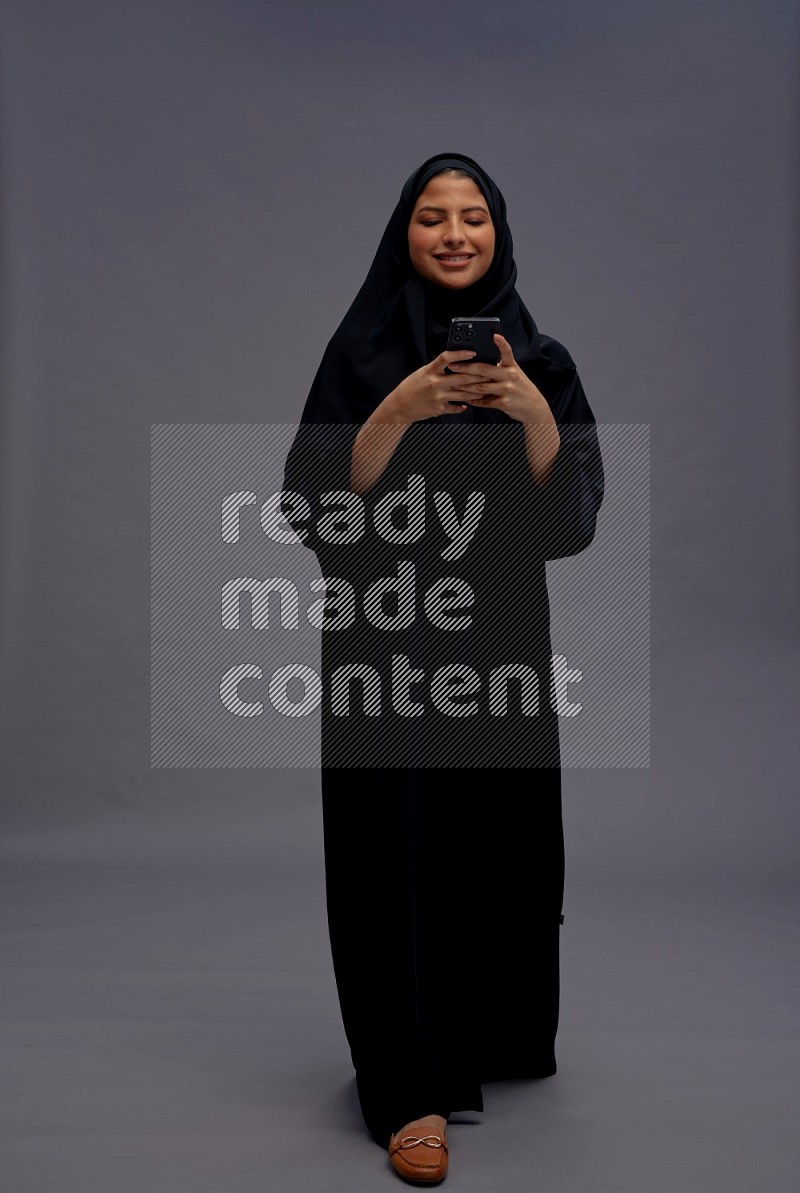 Saudi woman wearing Abaya standing texting on phone on gray background