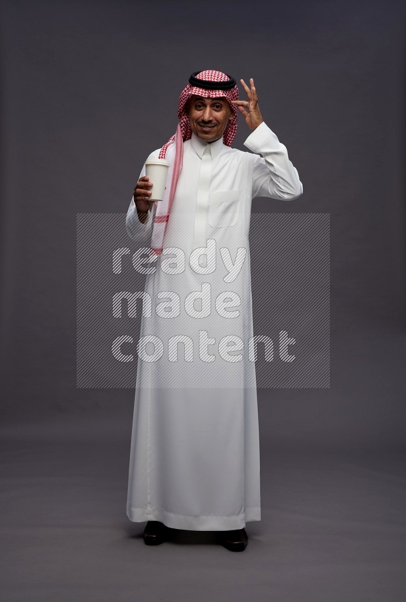 Saudi man wearing thob and shomag standing holding paper cup on gray background