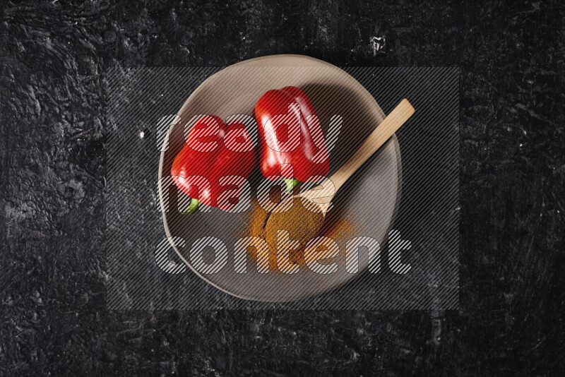 A wooden spoon full of ground paprika powder with red bell peppers, all on a pottery plate on black background