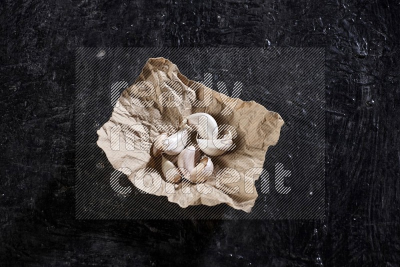 A crumpled piece of paper full of garlic cloves on a textured black flooring in different angles