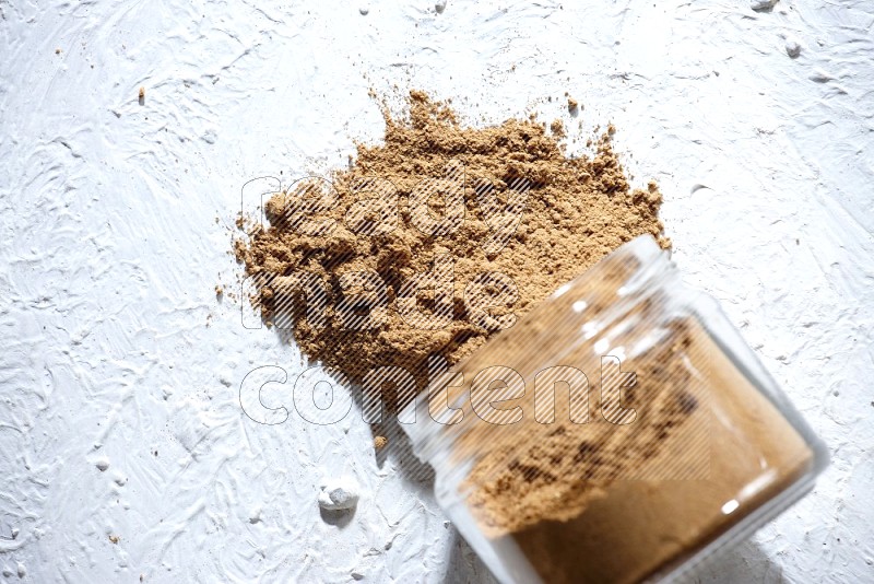 A flipped glass jar full of allspice powder and powder spilled out of it on a textured white flooring