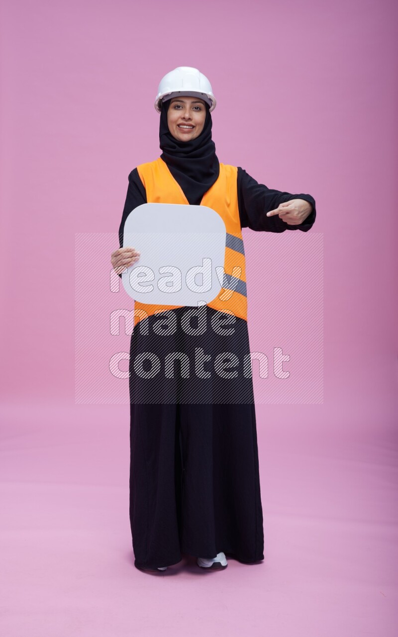 Saudi woman wearing Abaya with engineer vest and helmet standing holding social media sign on pink background