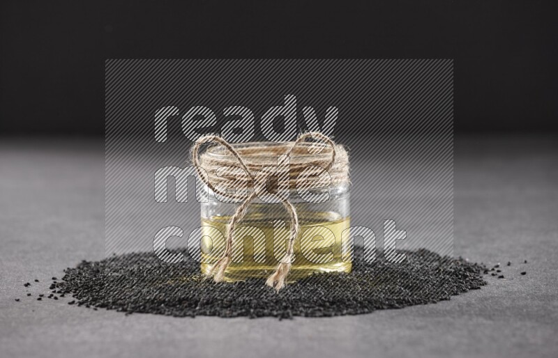 A glass jar full of black seeds oil surrounded by the seeds on a black flooring