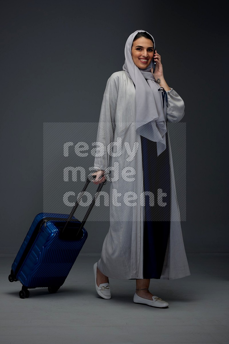 A Saudi woman wearing a light gray Abaya and head scarf standing and talking in the phone while holding shopping bags eye level on a grey background