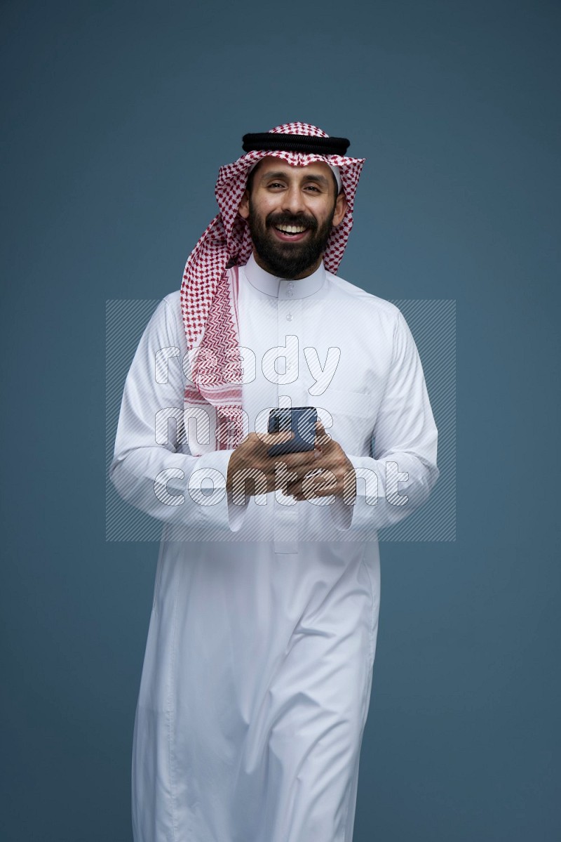 A Male posing with a phone in a blue background wearing Saudi Thob and Shomag