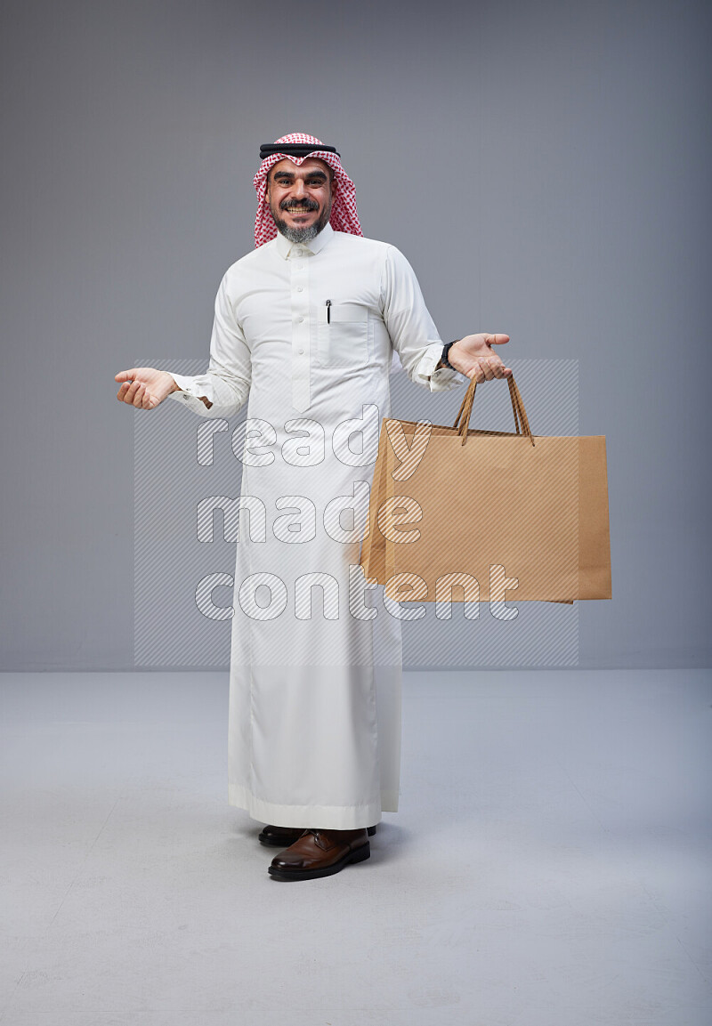 Saudi man Wearing Thob and red Shomag standing holding shopping bag on Gray background