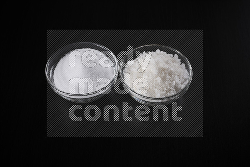 2 glass bowls one is filled with table salt and the other with coarse sea salt on black background