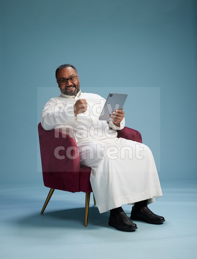 Saudi Man without shimag sitting on chair working on tablet on blue background