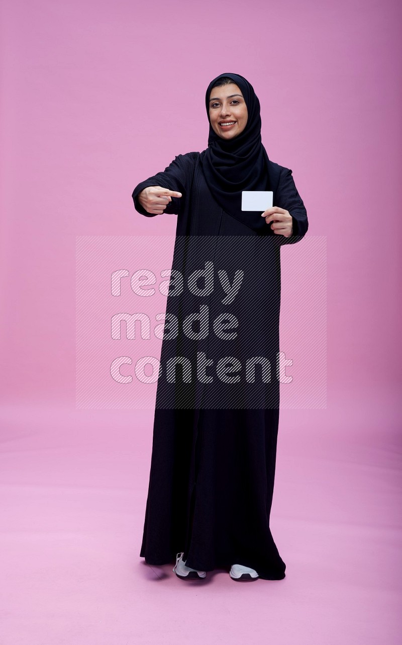 Saudi woman wearing Abaya standing holding ATM card on pink background