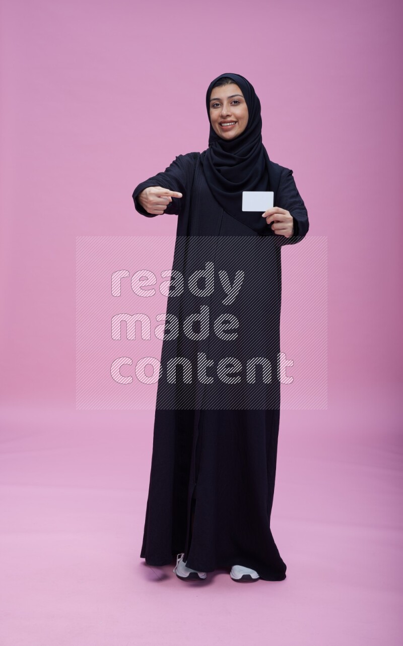 Saudi woman wearing Abaya standing holding ATM card on pink background