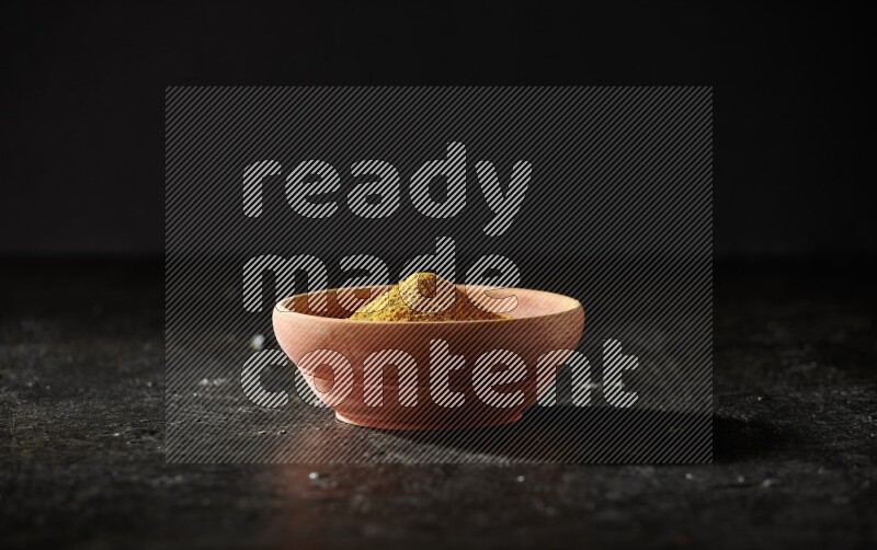 A wooden bowl full of turmeric powder with dried turmeric fingers on textured black flooring
