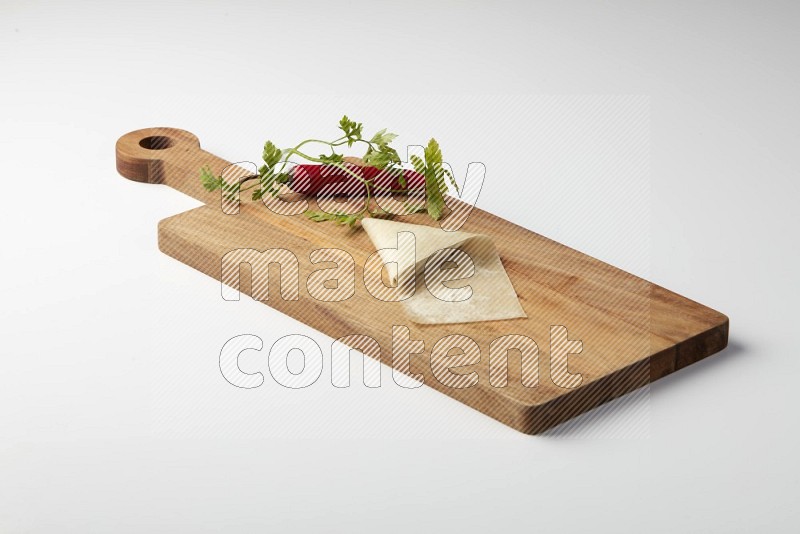 One uncooked samosa with a single red pepper and parsley on a wooden cutter on a white background