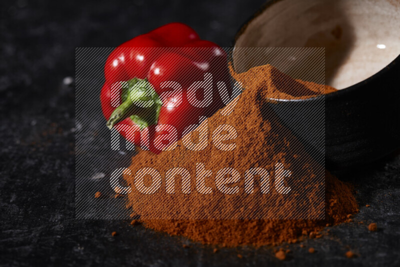 A black pottery bowl full of ground paprika powder with fallen powder from it on black background