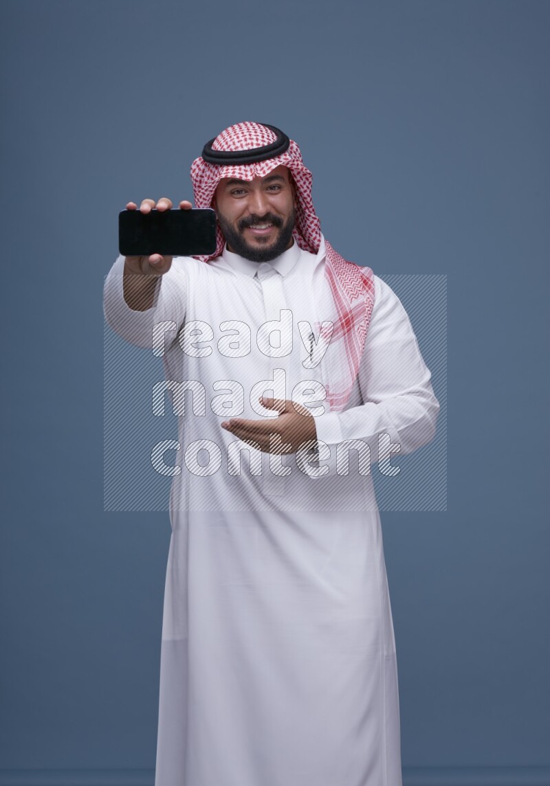 A man Showing his smartphone Screen on Blue Background wearing Saudi Thob