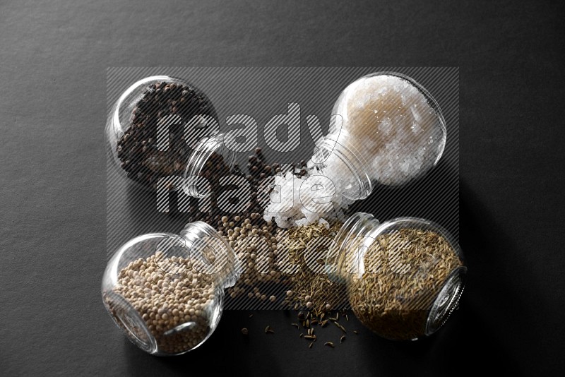 4 glass spice jars full of salt, black peppers, white peppers and cumin on black flooring