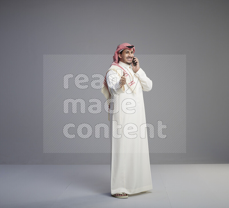 A Saudi man standing wearing thob and red shomag talking on phone on gray background