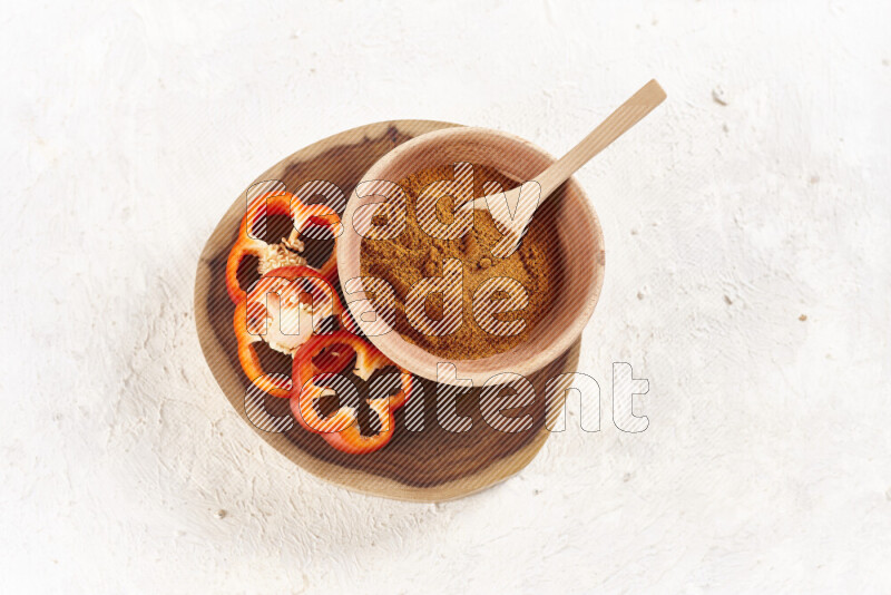 A wooden bowl full of ground paprika powder and sliced red bell pepper beside it all on a wooden tray on white background