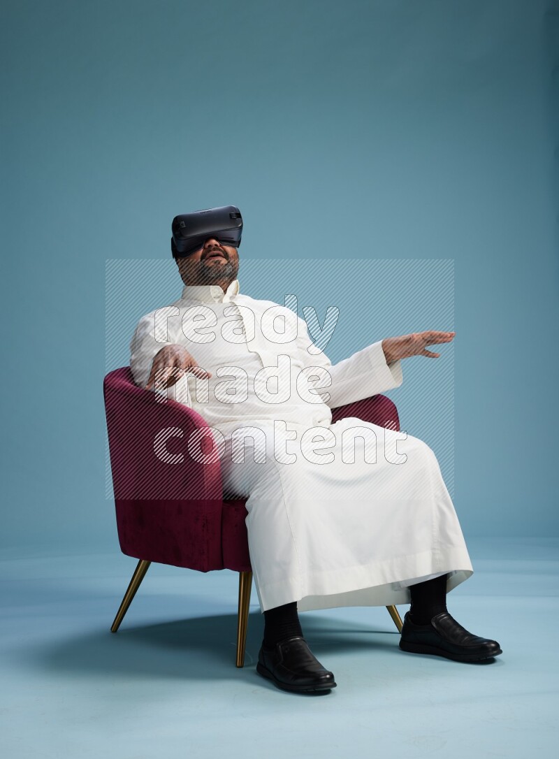 Saudi Man without shimag sitting on chair  gaming with VR on blue background