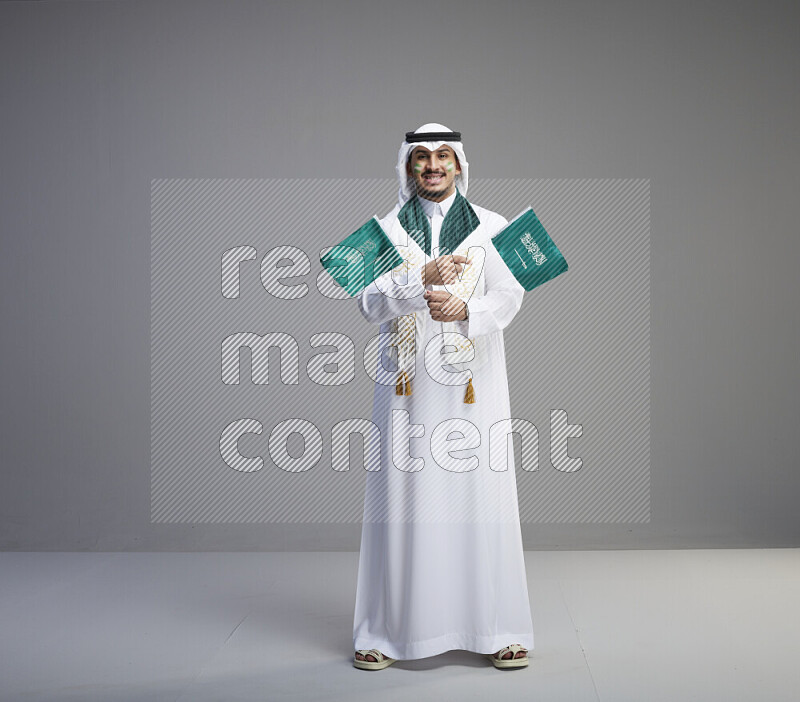A saudi man standing wearing thob and white shomag with face painting and saudi flag scarf and holding small saudi flag on gray background