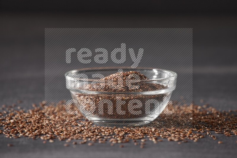 A glass bowl full of flaxseeds surrounded by the seeds on a black flooring
