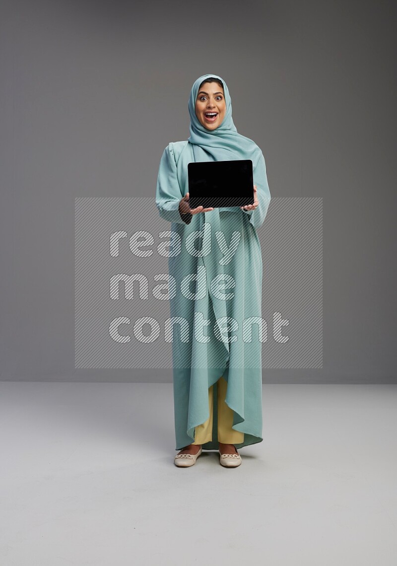 Saudi Woman wearing Abaya standing showing tablet to camera on Gray background