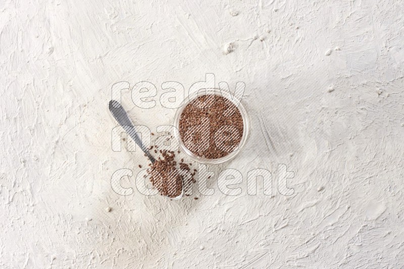 A glass jar full of flax and a metal spoon full of the seeds on a textured white flooring in different angles