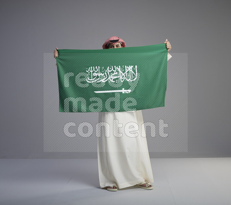 A saudi man standing wearing thob and red shomag holding big saudi flag on gray background