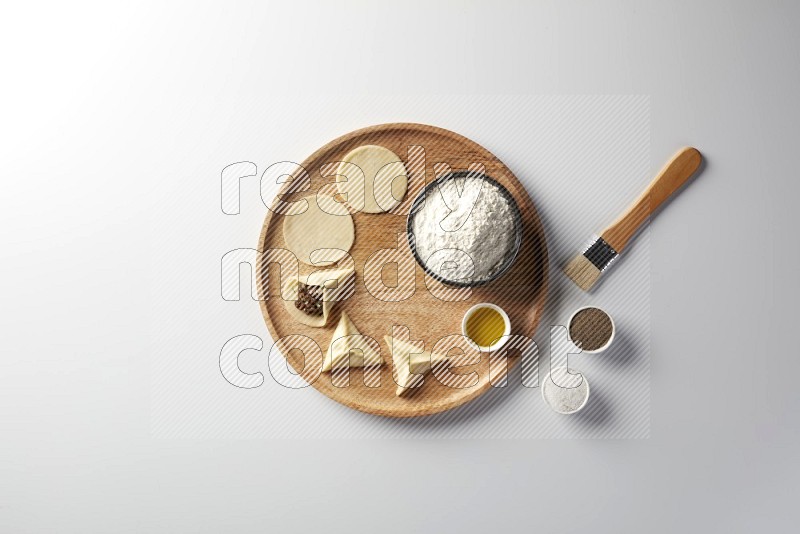 two closed sambosas and one open sambosa filled with meat while flour, salt, black pepper and oil with oil brush aside in a wooden dish on a white background