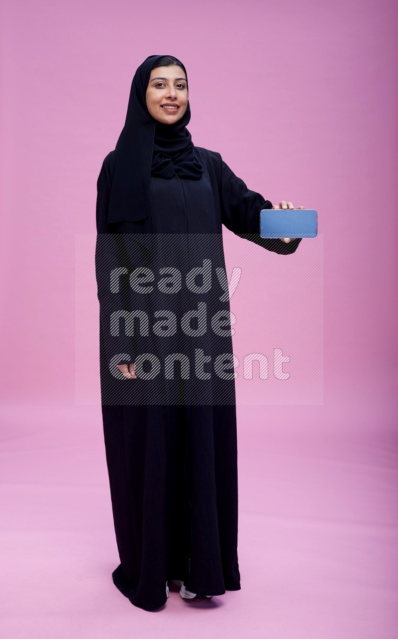 Saudi woman wearing Abaya standing showing phone to camera on pink background