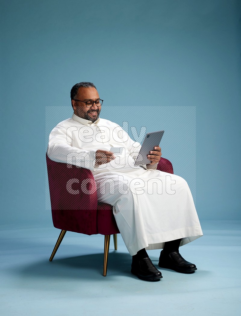 Saudi Man without shimag sitting on chair holding ATM card while working on tablet on blue background