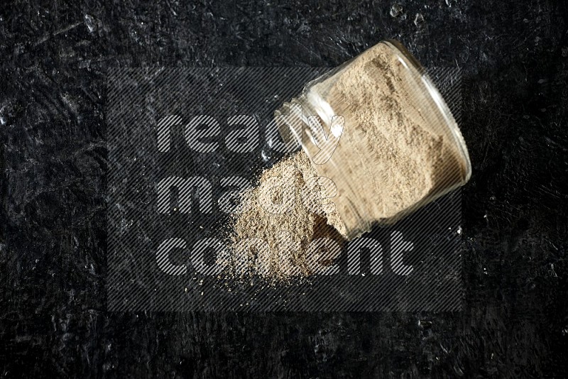 A flipped glass jar full of cardamom powder and powder spilled out of it on textured black flooring