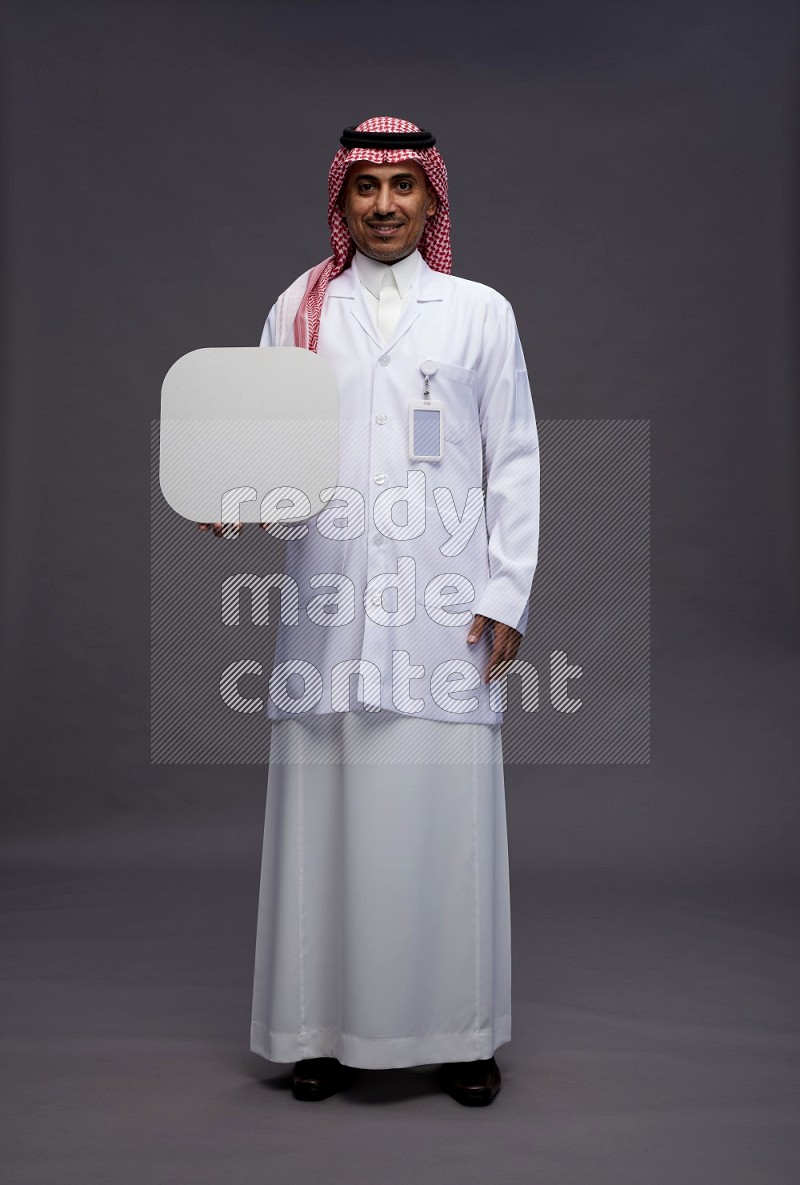 Saudi man wearing thob with lab coat and shomag with pocket employee badge standing holding social media sign on gray background