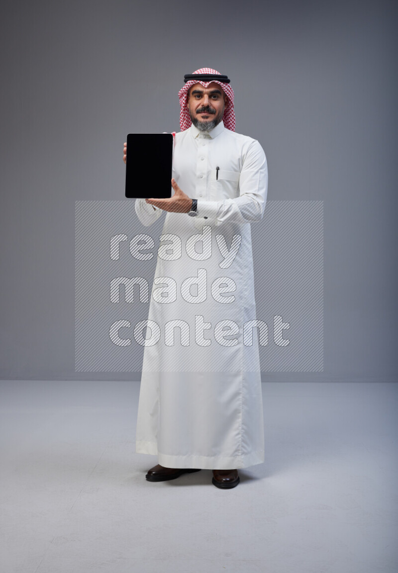 Saudi man Wearing Thob and red Shomag standing showing tablet to camera on Gray background