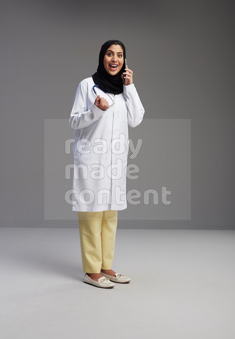 Saudi woman wearing lab coat with stethoscope standing talking on phone on Gray background