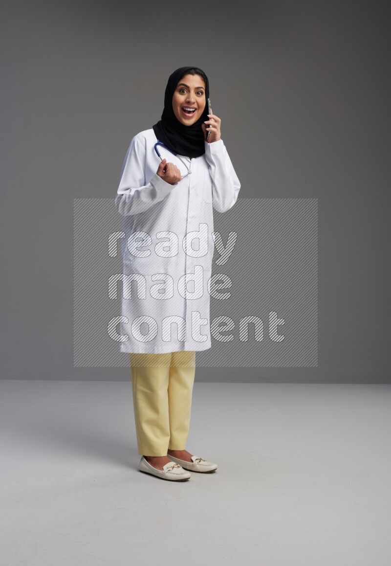 Saudi woman wearing lab coat with stethoscope standing talking on phone on Gray background