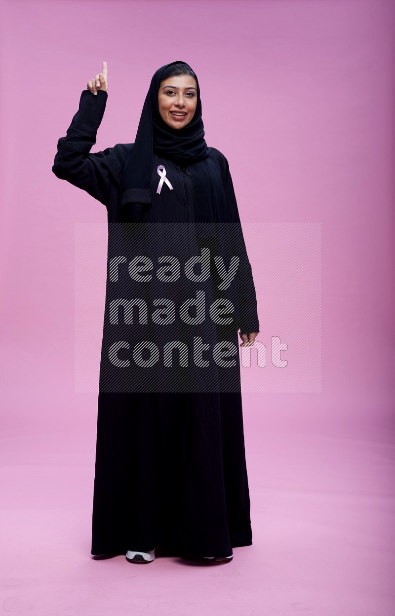 Saudi woman wearing pink ribbon on Abaya standing interacting with the camera on pink background