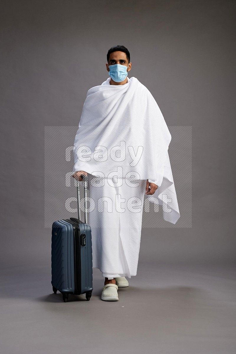 A man wearing Ehram with face mask Standing holding traveling bag on gray background