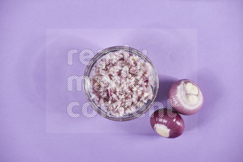 A glass bowl full of chopped red onions with halved onions beside it on purple background