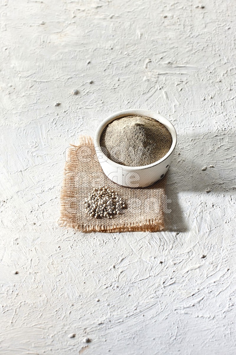 White pottery bowl full of white pepper powder set on a burlap piece of fabric with pepper beads and wooden pepper grinder on textured white flooring