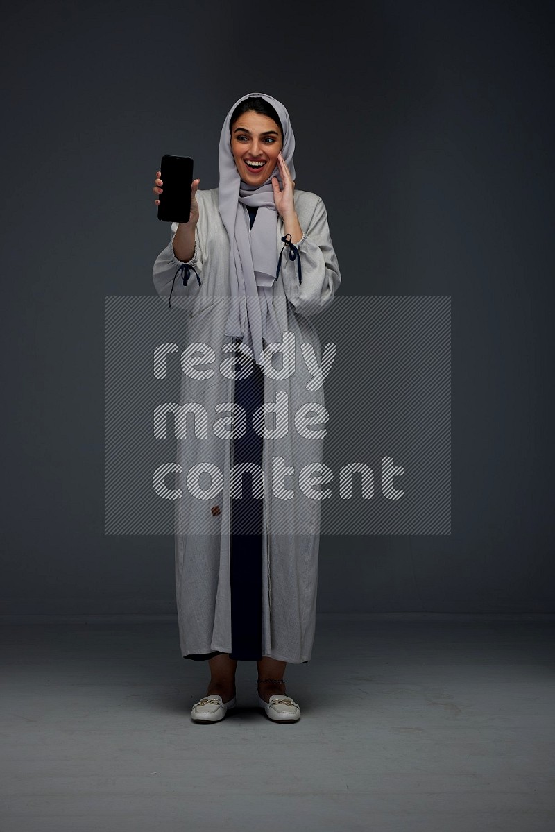 A Saudi woman wearing a light gray Abaya and head scarf standing and showing the phone's screen while holding a blue luggage eye level on a grey background