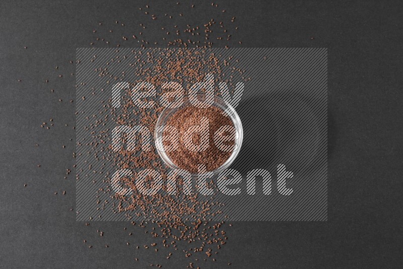 A glass bowl full of garden cress seeds surrounded by seeds on a black flooring