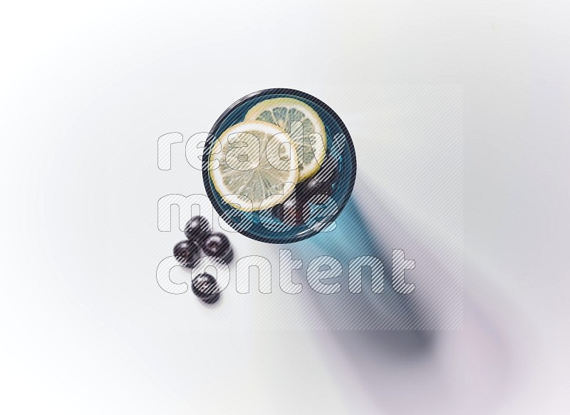 Blue transparent glass filled with blueberries decorated with lemon slices on a white background