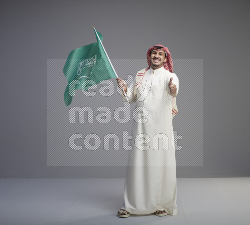 A Saudi man standing wearing thob and red shomag raising big Saudi flag on gray background