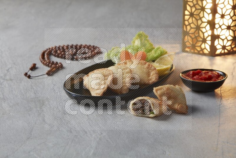 Three fried sambosas in an oval shaped black plate and a red sauce in a black round ramekin with a brown misbaha and a golden lantern on the side on a gray background